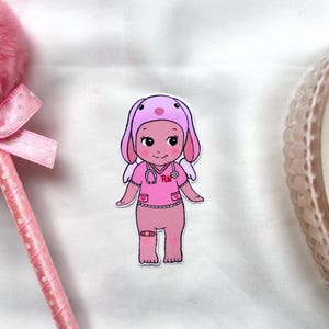 May include: A pink cartoon character wearing a pink bunny hat and a pink nurse uniform with the letters "RN" on the front. The character has pink wings and is holding a stethoscope.