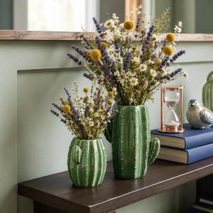 Op de afbeelding: Twee glanzende groene keramische vazen in de vorm van cactussen, elk voorzien van kleine witte stippen die stekels voorstellen. De vazen zijn gevuld met gedroogde bloemstukken van paarse lavendel, kleine witte madeliefjes en ronde gele craspedia.