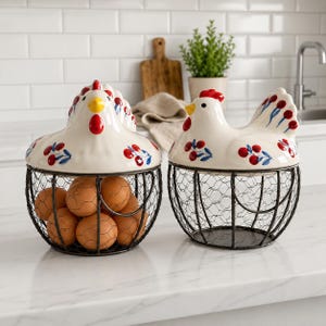 Op de afbeelding: Twee eierkorven van gaas met keramische deksels in de vorm van witte kippen. De deksels zijn voorzien van handgeschilderde rode bessen en blauwe bladeren, met gele snavels en rode kammen. Eén mand is gevuld met bruine eieren.