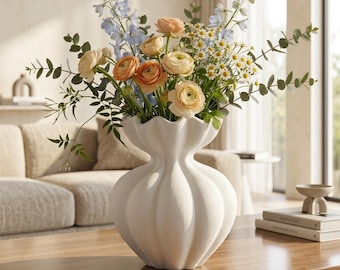 May include: A white ceramic vase with a scalloped, rounded shape holds a floral arrangement featuring light blue delphiniums, peach-colored ranunculus, white daisies, and green eucalyptus stems, creating a textured and layered botanical display.