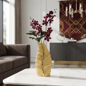 May include: A gold-colored vase shaped like a large, textured leaf, containing several stems of deep purple orchid flowers with green leaves. The vase has a prominent central vein and a metallic finish, standing upright on a flat surface.
