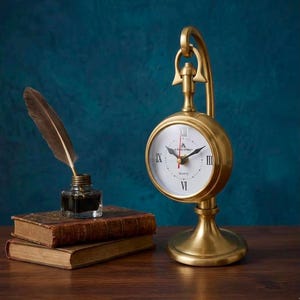 May include: A brass-toned metal desk clock with a white face, black Roman numerals, and a red second hand, suspended from a curved stand. Beside it are two stacked antique-style books and a glass inkwell containing a brown quill pen.