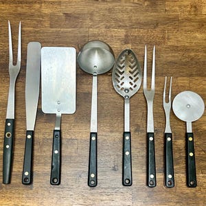 May include: A collection of stainless steel kitchen utensils with black handles. The set includes a spatula, slotted spoon, ladle, serving forks, and a pizza cutter. The handles are attached with brass rivets, and the tools are arranged on a wooden surface.