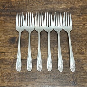 May include: A set of six silver-toned metal forks arranged in a row. Each fork features a slender handle with a decorative floral motif embossed at the base. The tines are long and pointed, showing a polished finish consistent across all pieces.