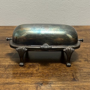 May include: A silver-toned metal butter dish with a domed lid and a rectangular base. The dish features four ornate, shell-shaped feet and a matching shell-shaped handle on the front. The metal surface shows signs of natural oxidation and patina.