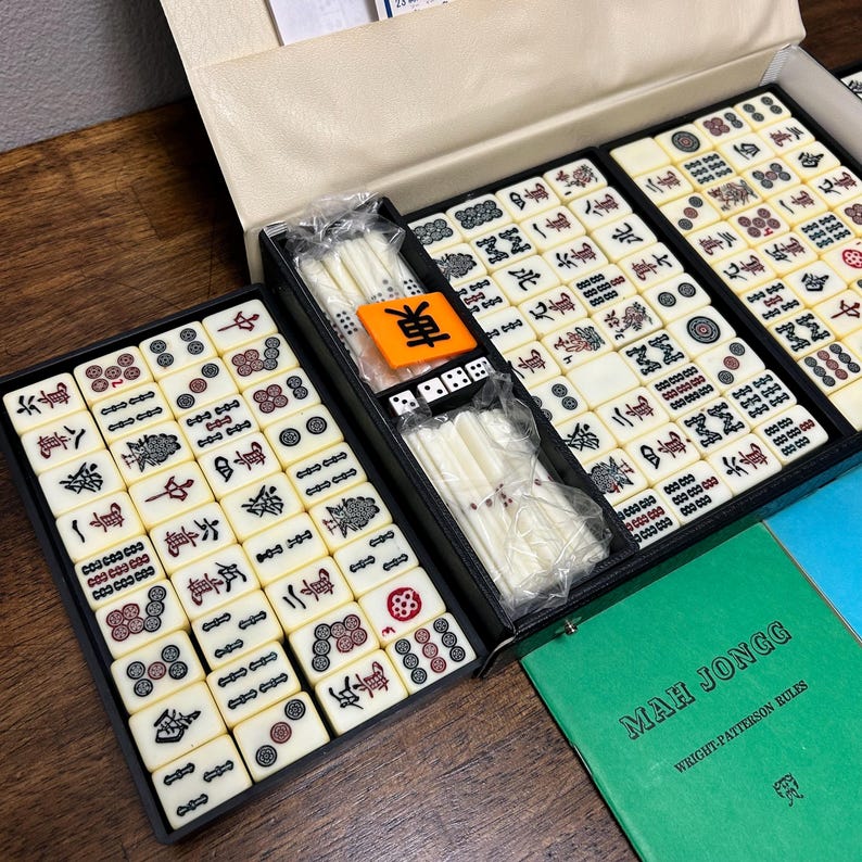 May include: A vintage Mahjong set with ivory-colored tiles, featuring red and black symbols and characters. The set includes dice, an orange tile, and a green booklet with the title "MAH JONGG". The tiles are arranged in black trays within a beige case.