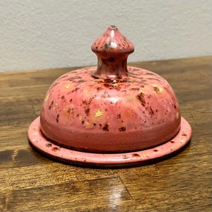 May include: A round ceramic butter dish with a dome-shaped lid featuring a pointed handle. The piece has a glossy pink glaze with mottled dark brown and gold-toned speckles throughout, and a smooth, rounded base plate with matching finish.