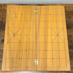 May include: A wooden folding game board with a grid pattern and black lines. The board is divided into two hinged sections, with brass-colored hinges. The board's surface has a light brown color.