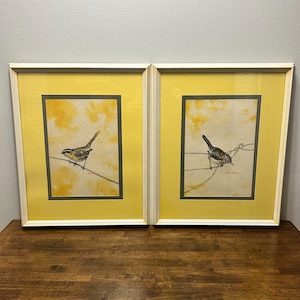 May include: Two framed prints featuring watercolor illustrations of small birds perched on thin branches. Each print has a bright yellow mat, a dark gray inner border, and a white frame. The background of the artwork is a soft yellow wash.