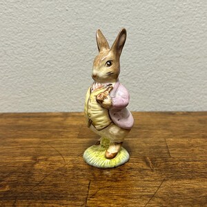 May include: A ceramic figurine of a rabbit wearing a pink jacket and yellow vest, holding a small object. The rabbit stands on a green and yellow base, with brown ears and a detailed face. The figurine is set against a neutral background.