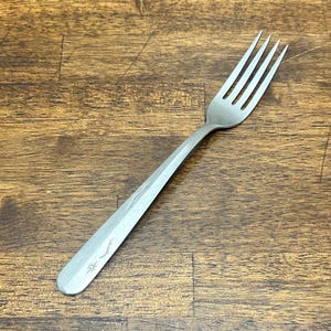 May include: A stainless steel dinner fork with a brushed matte finish. The handle features a subtle engraved pattern consisting of two small starburst motifs and two delicate wavy lines, providing a simple decorative detail on the flat surface.
