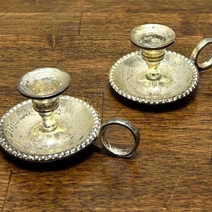 May include: Two silver-toned metal candle holders with a weathered, tarnished finish. Each holder features a circular base with a beaded edge, a central pedestal for a taper candle, and a small, circular side handle for carrying the object.