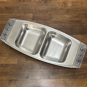 May include: A rectangular, divided serving dish made of brushed metal. The dish has two square compartments and decorative, dark-colored floral patterns on each end. The dish is designed for serving appetizers or snacks.