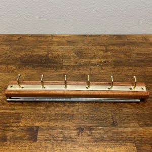 Könnte beinhalten: Eine Wandgarderobe mit einer horizontalen Holzbasis und einem mittigen, messingfarbenen Metallstreifen. Die Leiste verfügt über sechs gleichmäßig verteilte, nach oben gebogene messingfarbene Haken sowie sichtbare Montagelöcher an den Enden.