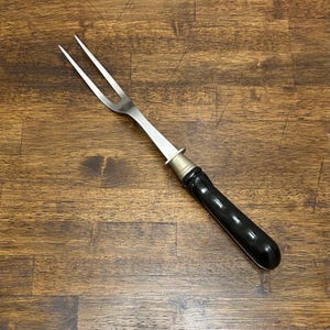 May include: A carving fork with a stainless steel head and a black handle. The fork has two tines and a silver ferrule where the handle meets the head. The handle is curved and glossy.