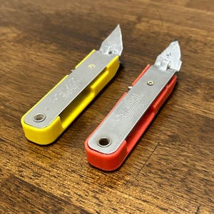 May include: Two Tap Boy brand can openers with metal blades and plastic handles, one in bright yellow and one in vibrant red. Each tool features a rectangular handle with a circular hole at the base and a pointed metal tip for piercing cans.