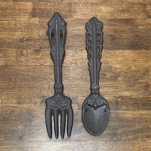 May include: A pair of dark brown cast iron wall decor pieces shaped like a large fork and a large spoon. Each utensil features an elongated handle with intricate, symmetrical scrollwork patterns and a solid, textured finish throughout the metal.