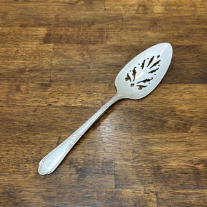 Puede incluir: Servidor de pastel plateado con un diseño calado decorativo en la hoja. El mango tiene un patrón floral en relieve. El servidor está hecho de un metal brillante y reflectante y descansa sobre una superficie de madera oscura.