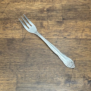 Könnte beinhalten: Eine silberfarbene Gabel mit drei Zinken, die ein dekoratives Blumenmuster am Griff aufweist. Das Besteck liegt diagonal auf einer dunklen Holzmaserung, was sein elegantes Design hervorhebt. Die Gabel ist aus Metall.