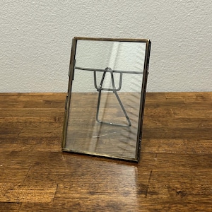 May include: A rectangular brass-toned metal and glass picture frame. The frame has clear glass front and back, with a metal stand. Designed to hold a photograph or artwork. The frame is displayed on a wooden surface.
