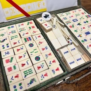 May include: A complete Mahjong set in a green case. The ivory-colored tiles feature red, green, and blue symbols and characters. The set includes dice, joker tiles, and a circular marker. The case is open, revealing the organized arrangement of the tiles.