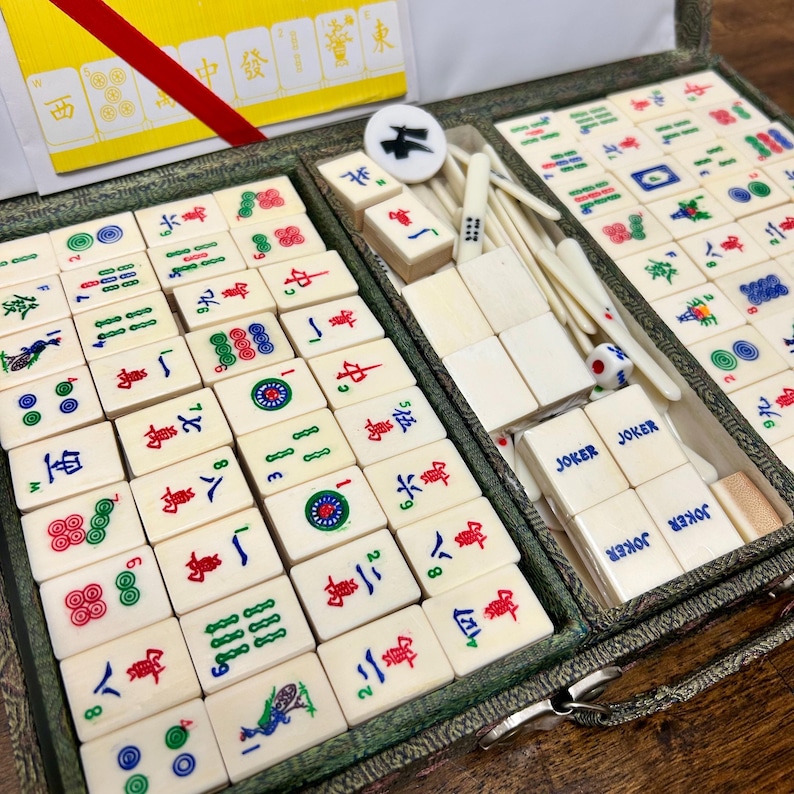May include: A complete Mahjong set in a green case. The ivory-colored tiles feature red, green, and blue symbols and characters. The set includes dice, joker tiles, and a circular marker. The case is open, revealing the organized arrangement of the tiles.