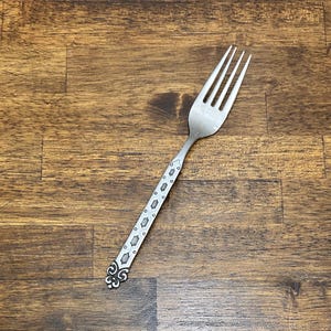 Puede incluir: Un tenedor plateado con cuatro púas y un mango decorativo. El mango presenta un patrón repetitivo de pequeños óvalos y diamantes, con un diseño ornamentado en el extremo. El tenedor es de metal brillante.