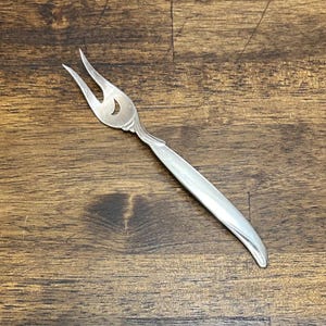 May include: A silver-toned metal cocktail fork featuring two slender, curved tines and a smooth, tapered handle. The base of the tines displays a small, crescent-shaped cutout detail, and the overall design has a polished, reflective finish.