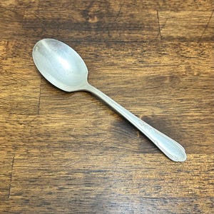 May include: A silver spoon with a rounded bowl and a decorative handle. The spoon is made of a shiny metal and is resting on a wooden surface. The handle has a floral design.