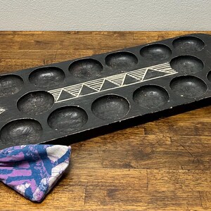 Op de afbeelding: Een zwart houten Mancala-bord met 12 ronde inkepingen en twee handvatten. Het bord heeft witte geometrische ontwerpen. Een klein trekkoordzakje met een blauw, paars en wit patroon ligt ernaast.