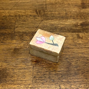 May include: A small, rectangular trinket box with a floral design on the lid. The box is made of a light-colored stone, possibly marble or onyx. The floral design features pink and white petals and a green leaf.