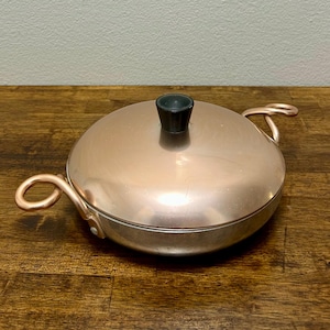 Op de afbeelding: Een ronde, koperkleurige pan met een bijpassend deksel en twee gebogen handgrepen aan de zijkanten. Het deksel is voorzien van een kleine, zwarte, cilindervormige knop in het midden. De pan heeft een metallic afwerking en de grepen zijn geklonken.