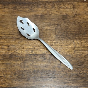 Op de afbeelding: Een zilverkleurige serveerlepel met een blad-vormige kop en decoratieve uitsparingen. De steel heeft een eenvoudig, elegant ontwerp. De lepel is van metaal en ligt op een donker houten oppervlak.