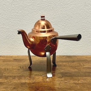 May include: A polished copper teapot featuring a rounded body, a curved spout, and a domed lid with a small finial. The teapot is supported by three slender, metallic legs and includes a long, straight, silver-toned handle extending from the side.
