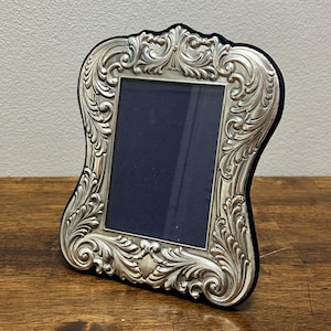May include: Ornate silver-toned picture frame with a dark blue interior. The frame features detailed scrollwork and a curved, elegant shape. The frame is displayed on a wooden surface.