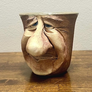 May include: A ceramic mug featuring a sculpted, three-dimensional human face with a large nose, heavy eyelids, and blue eyes. The mug has a textured, earthy brown finish with lighter beige highlights across the facial features.