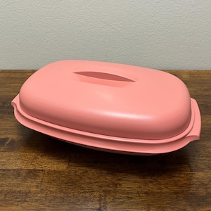 Puede incluir: Un recipiente de plástico rosa vintage con tapa. El recipiente tiene forma rectangular con bordes redondeados y un asa a cada lado. La tapa tiene una abertura larga y ovalada en el centro.
