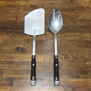 Puede incluir: Un juego de espátula y cuchara de acero inoxidable. La espátula tiene una hoja plana y en ángulo, mientras que la cuchara tiene un cuenco redondeado. Ambos tienen mangos largos de metal con empuñaduras de plástico negro y remaches dorados. El juego está diseñado para cocinar.