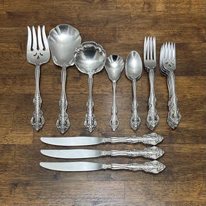 May include: A collection of ornate silver-colored flatware, including forks, spoons, and knives, arranged on a wooden surface. The silverware features detailed floral and scrollwork patterns on the handles. The set includes serving spoons and multiple forks and knives.