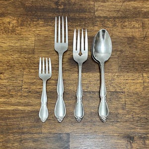 May include: Four pieces of vintage silverware are arranged on a wooden surface. The set includes a small fork, a large fork, a fork with a decorative cutout, and a spoon. The silverware has a silver finish and ornate detailing on the handles.