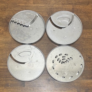 May include: Four stainless steel food processor blades on a wooden surface. The blades have different cutting designs, including slicing, grating, and shredding. The blades are circular and have various holes and slots for food preparation.
