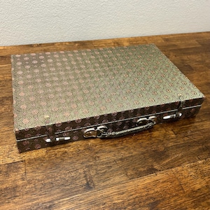 May include: A rectangular, patterned box with a handle and metal clasps. The box has a green background with a repeating pink floral design. The handle and clasps are silver-toned. The box is sitting on a wooden surface.