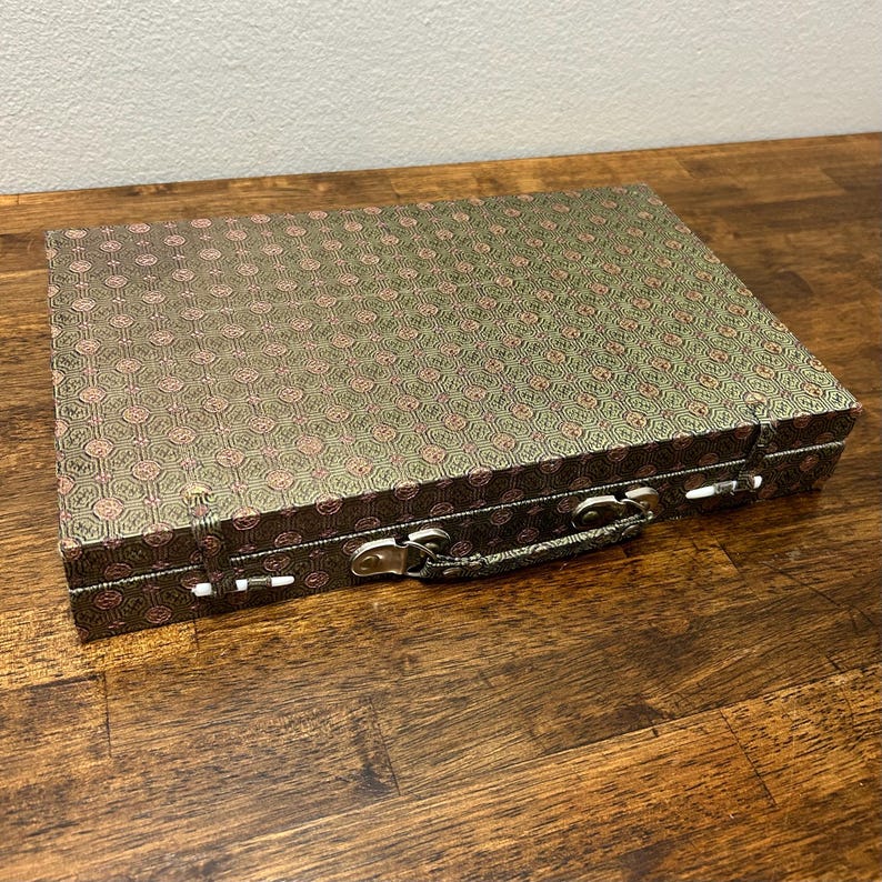 May include: A rectangular, patterned box with a handle and metal clasps. The box has a green background with a repeating pink floral design. The handle and clasps are silver-toned. The box is sitting on a wooden surface.