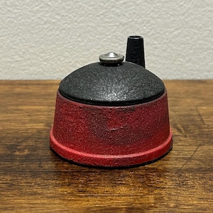 May include: A small, vintage-style container with a black, textured top and a red base. The container has a small, cylindrical black spout and a silver-colored knob on top. The base is a deep red color, with a slightly faded appearance.