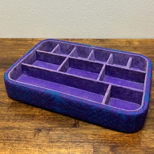 May include: A rectangular jewelry organizer with a patterned blue and purple exterior and a purple velvet interior. The organizer has multiple compartments of various sizes for storing jewelry. The box is sitting on a wooden surface.