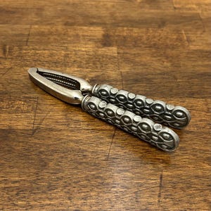 May include: A silver-colored nutcracker with ornate, textured handles. The nutcracker is made of metal and is designed to crack nuts. The handles have a repeating pattern of circles and ovals.