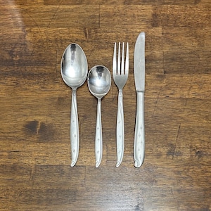 Puede incluir: Un juego de cuatro utensilios plateados: una cuchara, una cuchara más pequeña, un tenedor y un cuchillo. Cada pieza tiene un mango delgado con un diseño de estrella. Los utensilios están colocados sobre una superficie de madera.