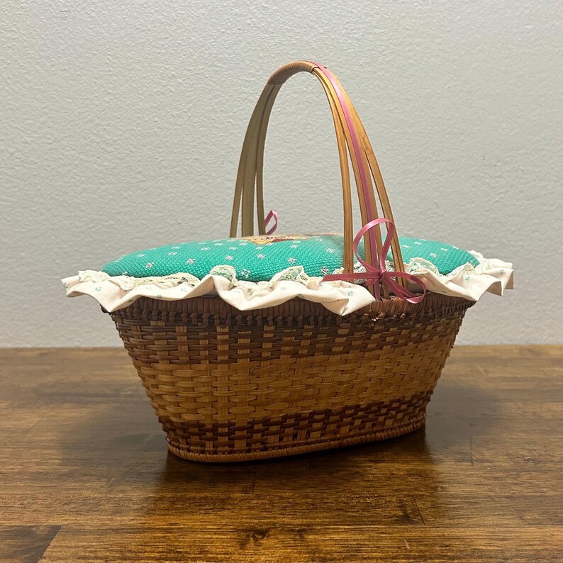 Puede incluir: Una cesta de mimbre marr&oacute;n tejida con una tapa de tela turquesa con un peque&ntilde;o estampado floral. La tapa est&aacute; bordeada con un volante de tela color crema y acentuada con una cinta rosa atada al asa alta y arqueada de tiras de madera.