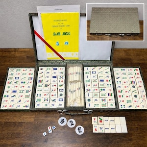 May include: Open Mah Jong set with tiles, dice, and counters. The set includes a yellow instruction booklet with the title "STANDARD RULES for the CHINESE DOMINO GAME of MAH JONG". The tiles are white with various symbols and characters. The case is green.