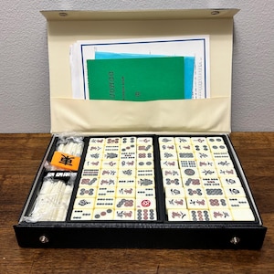 May include: A black Mahjong set with ivory tiles, dice, and a wind indicator. The set includes a green booklet labeled "JONGG PATTERSON RULES" and is stored in a black case with a beige interior. The tiles feature Chinese characters and symbols.